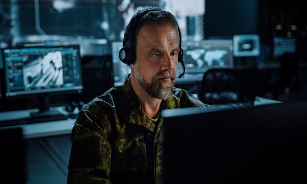 Bearded Military Surveillance Officer in Headset Working in a Central Office Hub for Cyber Operations, Control and Monitoring for Managing National Security, Technology and Army Communications. Bearded Military Surveillance Officer in Headset Working in a Central Office Hub for Cyber Operations, Control and Monitoring for Managing National Security, Technology and Army Communications.